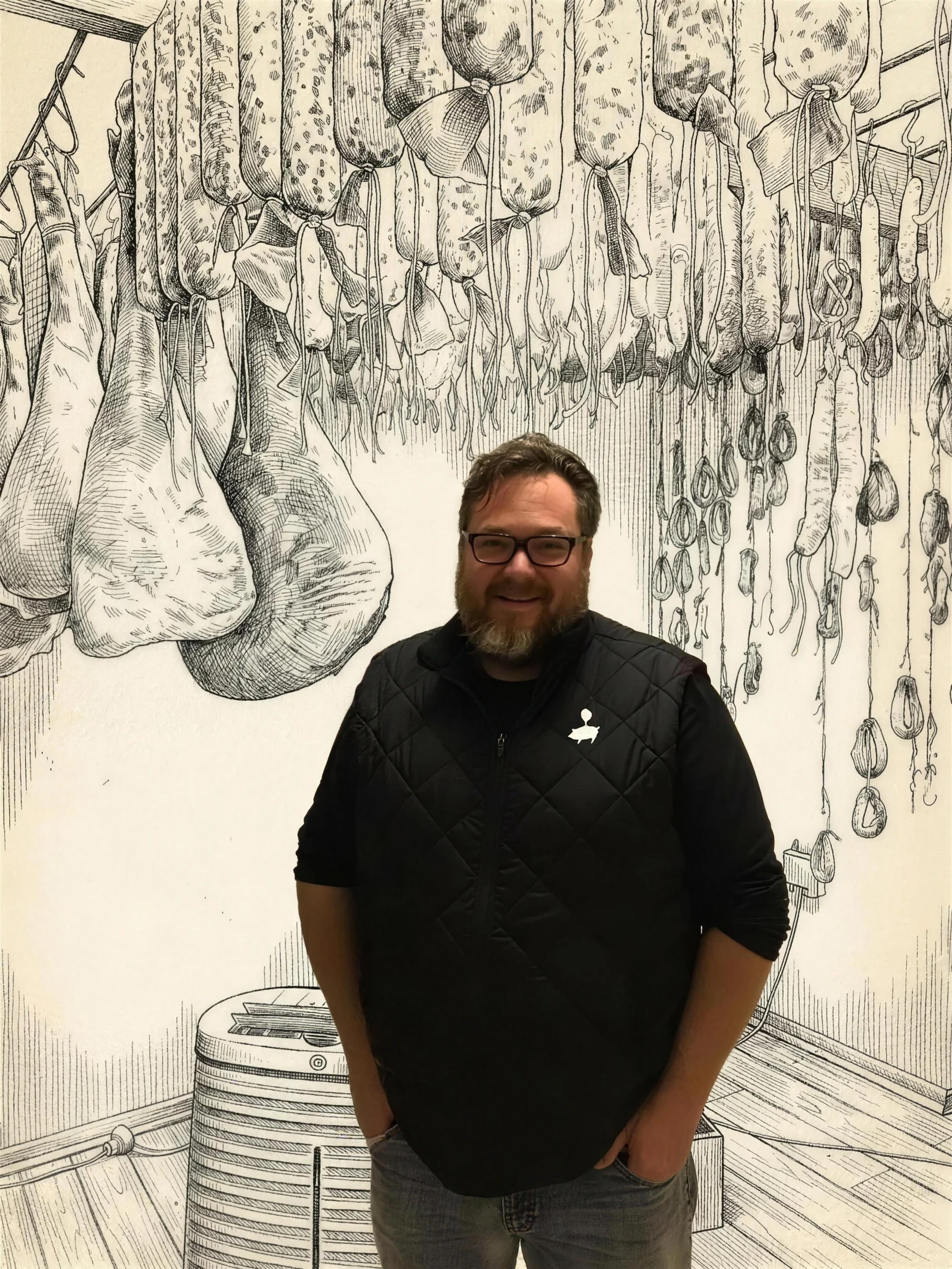 Gilbert Ferreira, photographed, standing in his maturing chamber beneath a hand-drawn canopy of bloomed salami and hams.